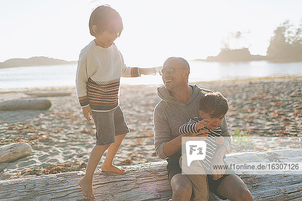 Lächelnder Vater mit Kindern auf Treibholz am Strand sitzend