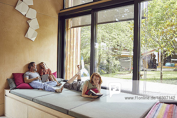 Frau  die ein Buch liest  während Vater und Tochter auf dem Bett zu Hause ein digitales Tablet benutzen