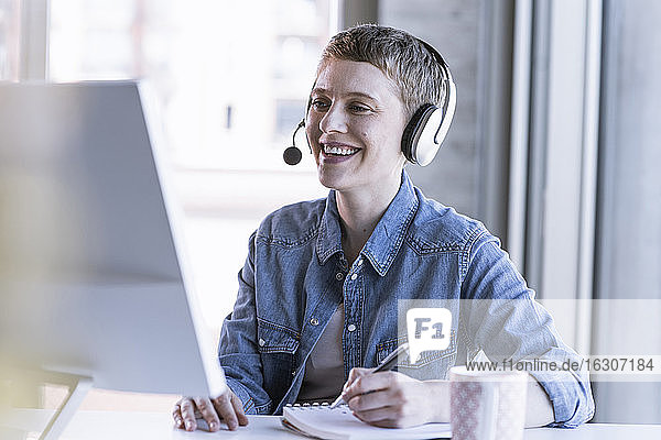 Lächelnde Geschäftsfrau mit Headset am Schreibtisch im Büro  die Notizen macht