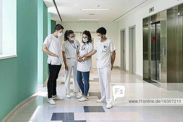 Ärzte  die chirurgische Masken tragen  diskutieren über ein digitales Tablet  während sie im Krankenhaus auf dem Flur stehen