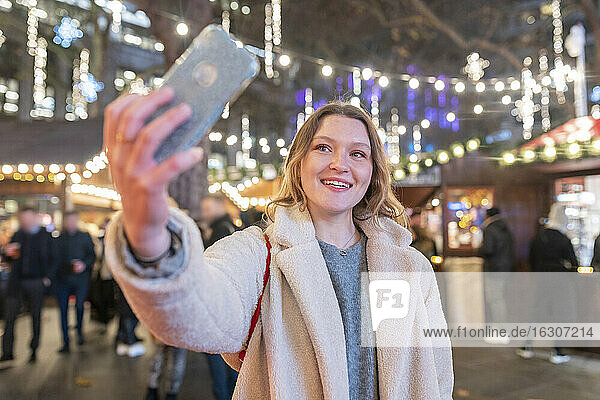 Junge Frau  die ein Selfie macht  während sie auf dem nächtlichen  beleuchteten Weihnachtsmarkt steht