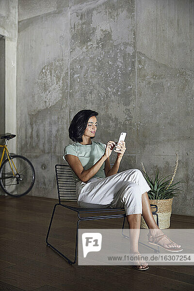 Geschäftsfrau  die ein Mobiltelefon benutzt  während sie auf einem Stuhl an der Wand im Büro sitzt