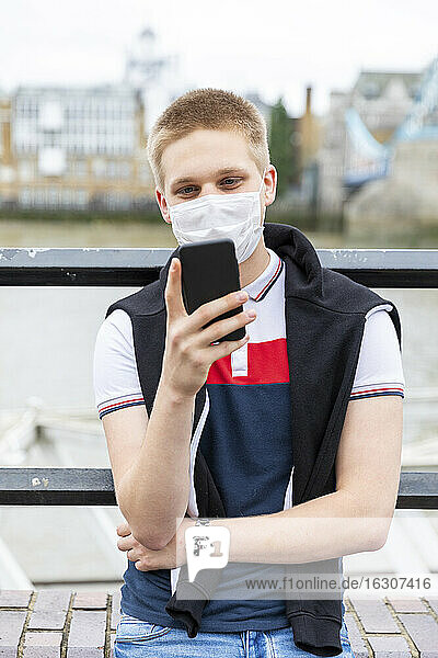 Junger Mann mit Gesichtsmaske bei der Nutzung eines Smartphones in einer Stadt während des Ausbruchs des Coronavirus