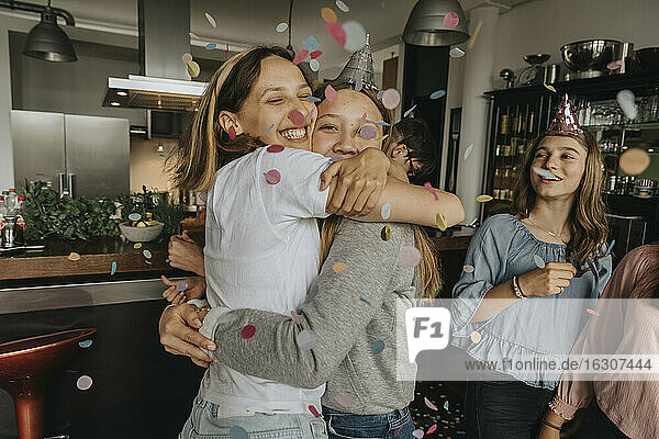 Glückliche Freunde  die sich umarmen  während sie inmitten von Konfetti auf einer Geburtstagsparty zu Hause stehen
