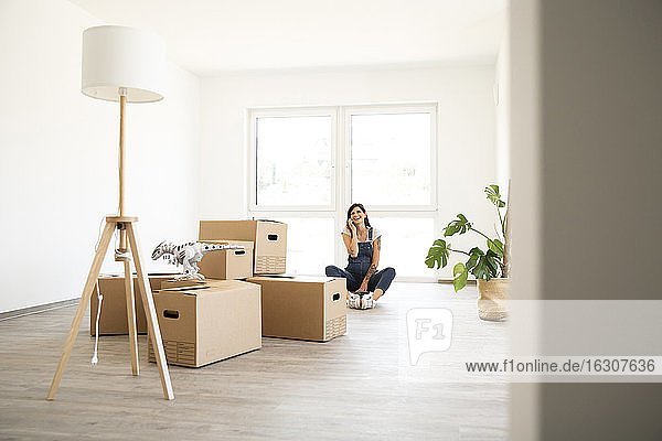 Pregnant woman talking over smart phone while sitting by cardboard boxes in new home