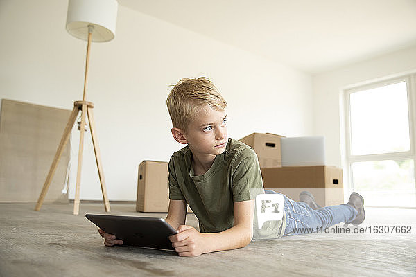 Junge benutzt digitales Tablet und schaut weg  während er in einem neuen Haus auf dem Boden liegt