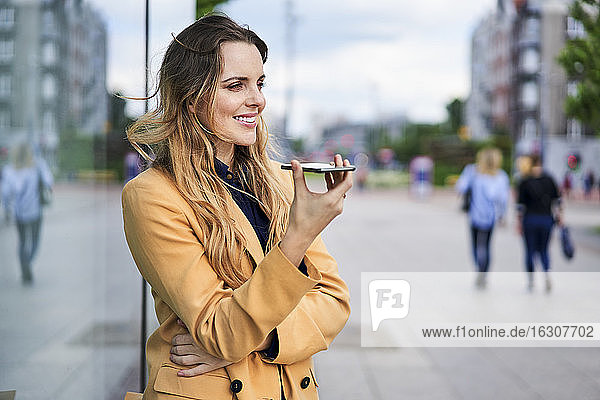 Lächelnde Frau  die an einer Straßenbahnhaltestelle einen Lautsprecher benutzt und über ihr Smartphone spricht