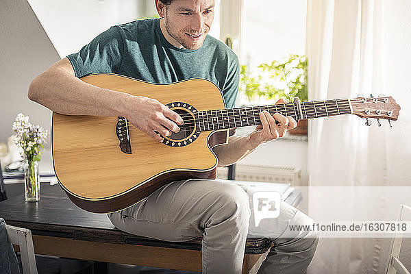 Lächelnder Mann spielt Gitarre  während er zu Hause auf dem Tisch sitzt