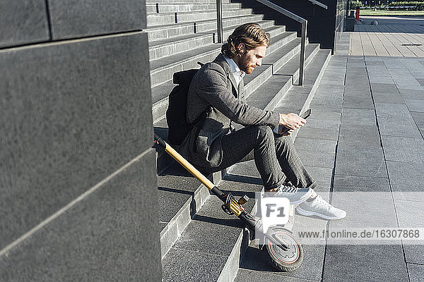 Männlicher Pendler  der ein digitales Tablet benutzt  während er mit einem Elektroroller auf einer Treppe im Stadtzentrum sitzt