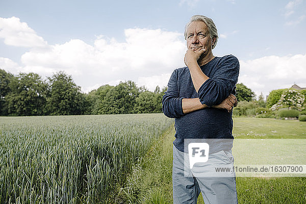 Wrinkled man looking away with hand on chin while standing against sky in field