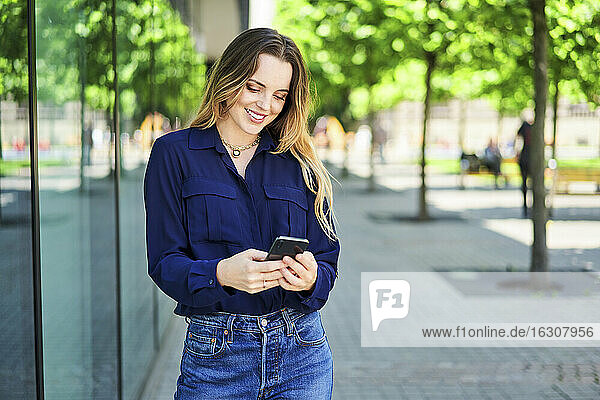 Lächelnde schöne Frau Text-Messaging durch Smartphone beim Stehen auf Gehweg in der Stadt