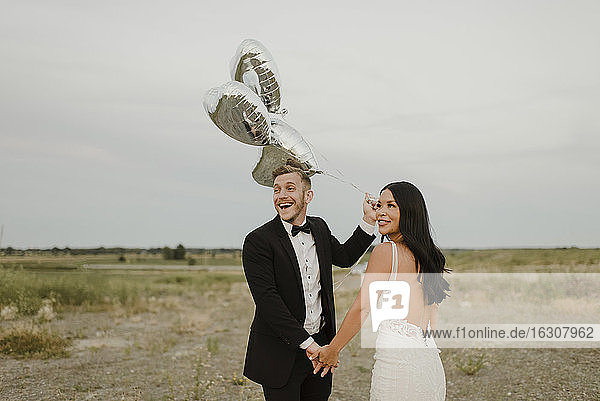 Glücklicher Bräutigam mit herzförmigen Luftballons hält die Hand der Braut gegen den Himmel
