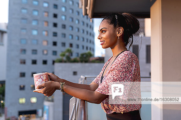 Schöne Frau hält Kaffeetasse  während sie auf dem Balkon steht und den Sonnenuntergang genießt