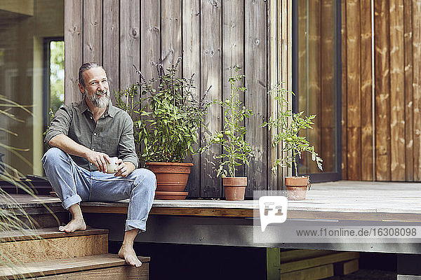 Smiling bearded man holding coffee cup while sitting outside tiny house