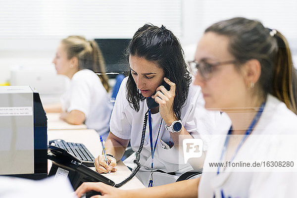 Female pharmacist writing while talking over telephone at desk in hospital
