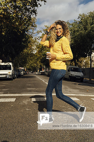 Lächelnde junge Frau beim Überqueren der Straße mit einem Einwegbecher in der Stadt an einem sonnigen Tag