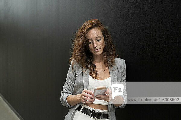 Geschäftsfrau  die ihr Smartphone benutzt und sich an eine schwarze Wand im Korridor lehnt