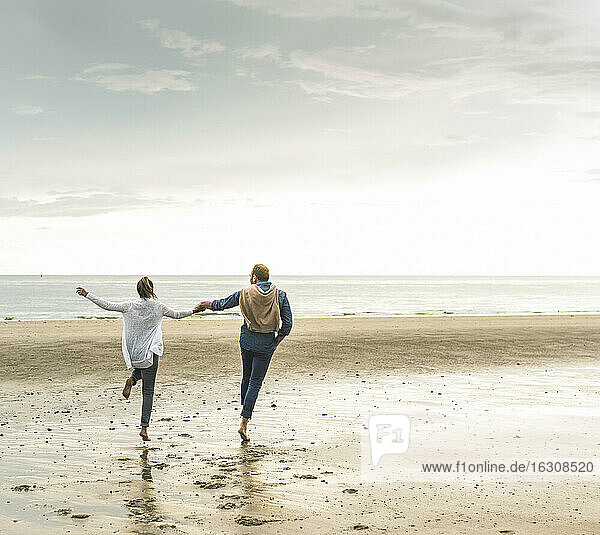 Cheerful mature couple holding hands while jumping at beach against cloudy sky