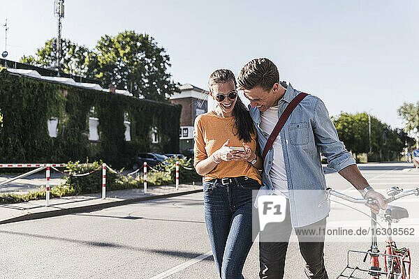 Fröhliches junges Paar schaut auf sein Smartphone  während es auf der Straße in der Stadt spazieren geht