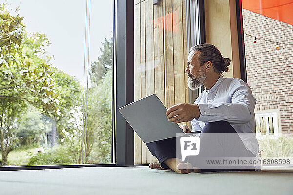 Bärtiger reifer Mann mit Laptop schaut durch das Fenster  während er in einem kleinen Haus sitzt