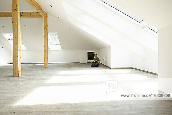 Junge spielt mit seinem Smartphone  während er auf dem Boden des Dachbodens eines neuen  unmöblierten Hauses sitzt