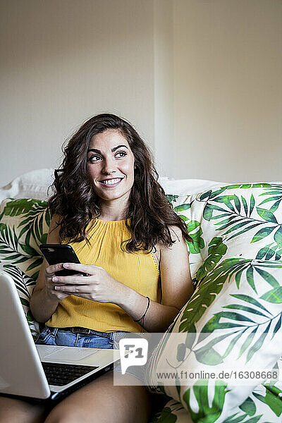 Thoughtful smiling woman holding mobile phone while sitting with laptop on armchair at home