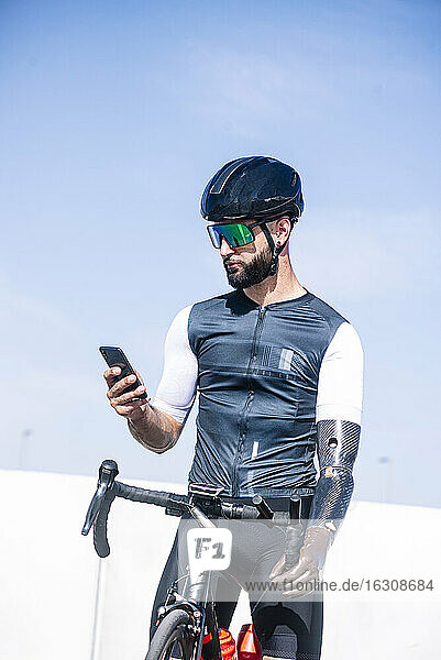 Männlicher amputierter Radfahrer  der sein Smartphone benutzt  während er mit dem Fahrrad vor einem klaren Himmel steht