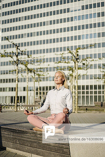 Weibliche Fachkraft meditiert im Lotussitz auf einer Bank vor einem Gebäude an einem sonnigen Tag