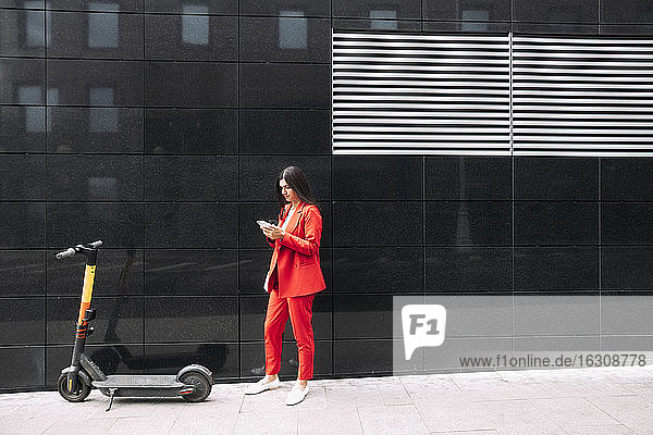 Unternehmerin in roter Geschäftskleidung  die ihr Smartphone benutzt  während sie vor einem schwarzen Gebäude in der Stadt steht
