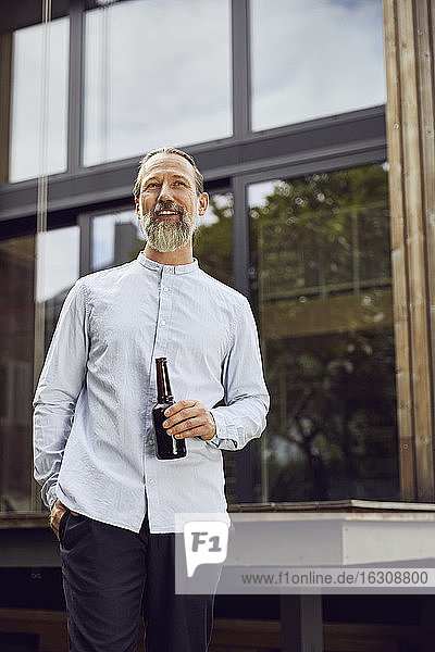 Bärtiger reifer Mann hält Bierflasche  während er vor einem kleinen Haus steht