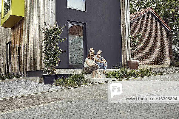 Eltern mit Tochter sitzen auf den Stufen vor einem kleinen Haus