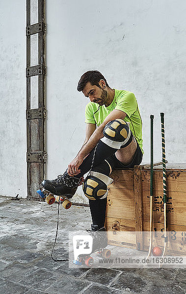 Mature male athlete wearing roller skate while sitting by hockey sticks on wooden box against wall at court