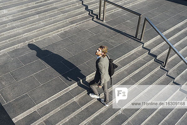 Männlicher Unternehmer mit Tasche  der eine Treppe hinaufgeht  während er in der Innenstadt an einem sonnigen Tag wegschaut
