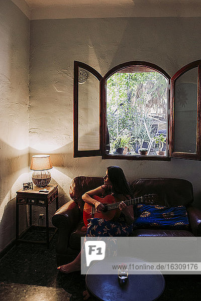 Junge Frau übt Gitarre  während sie auf dem Sofa im Wohnzimmer sitzt