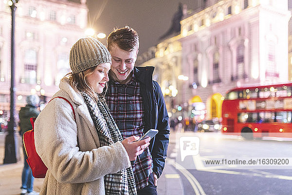 Frau benutzt Smartphone  während sie nachts mit ihrem Freund am Piccadilly Circus steht