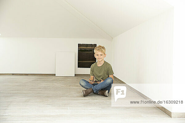 Lächelnder Junge  der sein Smartphone in der Hand hält  während er auf dem Boden im Dachgeschoss seines neuen Hauses sitzt