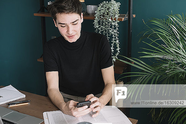 Gut aussehender junger Mann  der sein Smartphone benutzt  während er mit Büchern an einem Tisch vor einem Regal sitzt