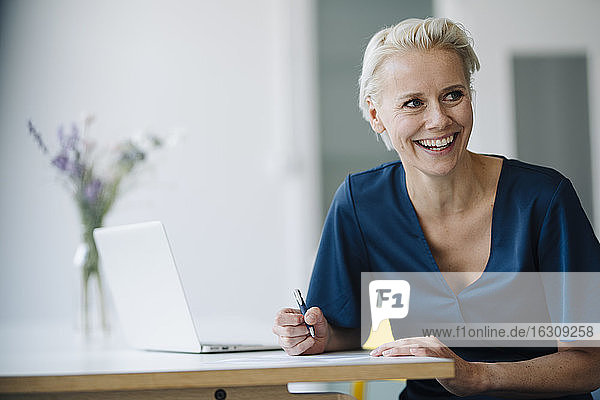 Fröhliche Geschäftsfrau mit Laptop auf dem Schreibtisch  die wegschaut  während sie im Büro sitzt