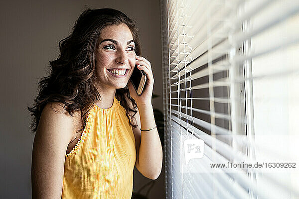 Fröhliche junge Frau  die mit ihrem Smartphone spricht  während sie durch die Jalousien zu Hause wegschaut