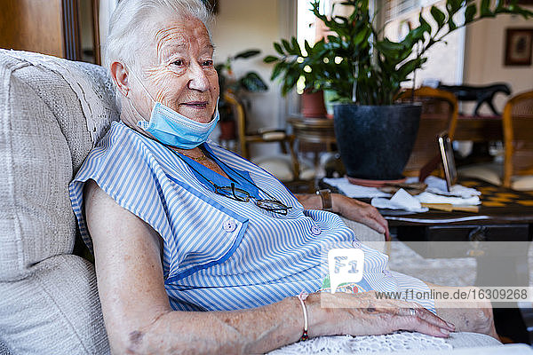 Weibliche Seniorin mit Schutzmaske sitzt zu Hause im Sessel