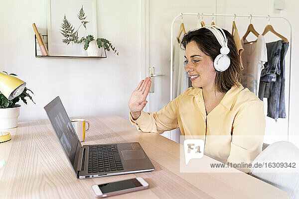 Lächelnde Frau winkt Freunden bei einem Videoanruf über den Laptop zu Hause zu