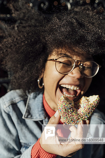 Nahaufnahme einer afroamerikanischen jungen Frau  die einen Donut hält und an einem sonnigen Tag im Freien lacht