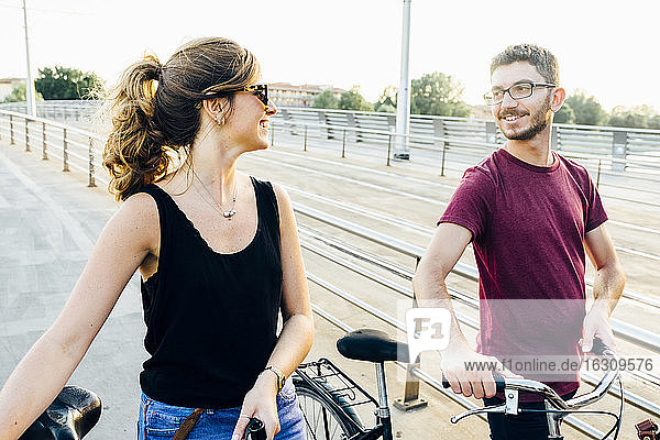 Couple talking while walking with bicycles on bridge against clear sky