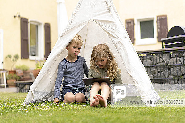 Niedliche Geschwister mit digitalem Tablet sitzen im Zelt auf dem Gras im Hinterhof gegen Haus