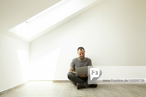 Älterer Mann  der einen Laptop benutzt  während er gegen eine weiße Wand im Dachgeschoss eines neuen Hauses sitzt