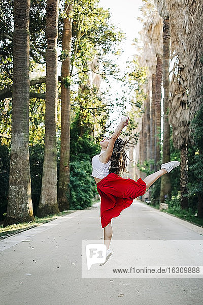 Junge Frau springt beim Tanzen auf der Straße zwischen Bäumen im Park
