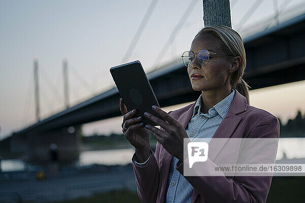 Selbstbewusste Unternehmerin vor einer Brücke in der Abenddämmerung mit digitalem Tablet