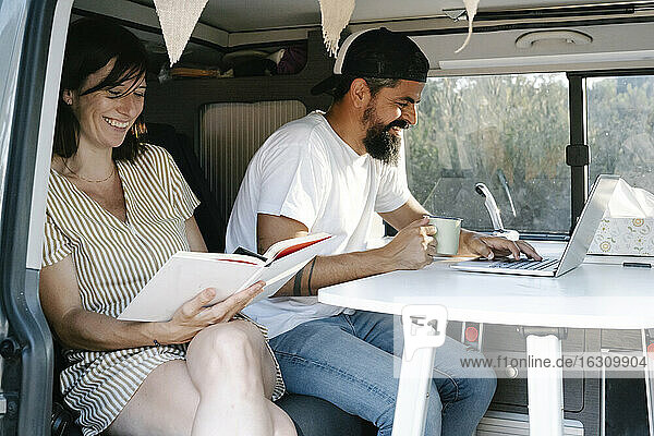 Lächelnde reife Frau  die ein Buch liest  während sie neben einem Mann sitzt  der einen Laptop im Lieferwagen benutzt