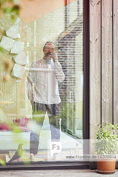 Mann spricht über Mobiltelefon  während er in einem kleinen Haus steht  das durch ein Fenster gesehen wird
