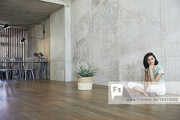 Nachdenkliche Geschäftsfrau sitzt mit Laptop auf Hartholzboden vor der Wand im Büro